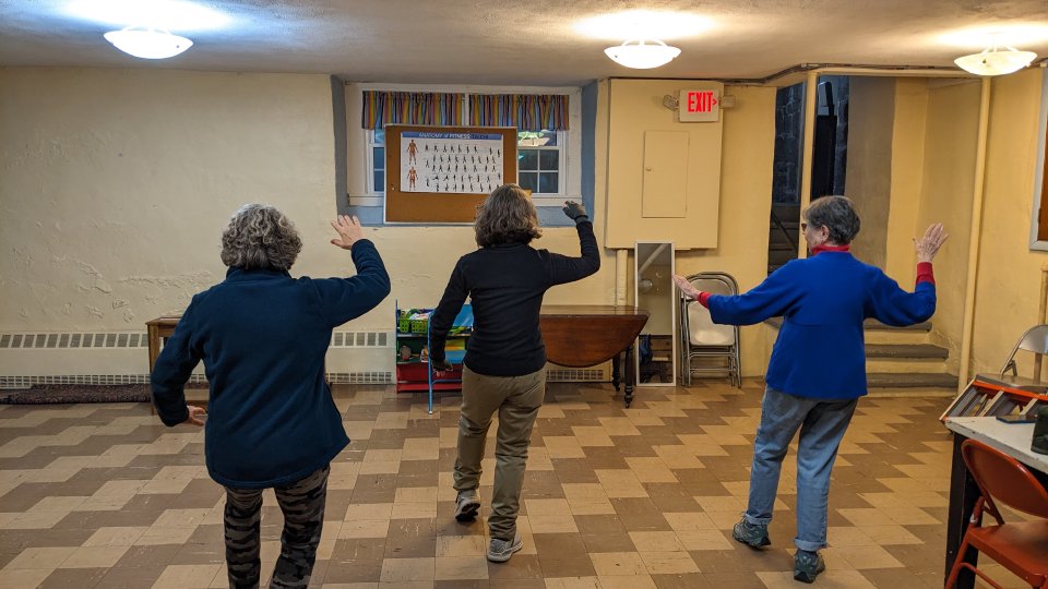 Three older adults in a bright, lively room joyfully lift arms together, connecting through group movement and community spirit.