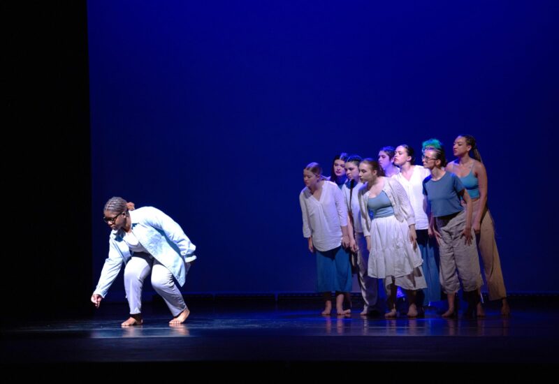 A lively group of Essex County dancers in casual wear unites on a blue-lit stage, one reaching down in a burst of creative energy.