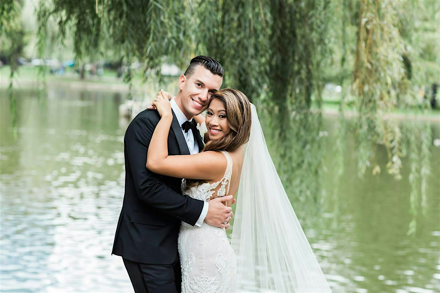 A joyful bride and groom in elegant wedding attire beam by an Essex County lake, celebrating love amid our vibrant community backdrop.