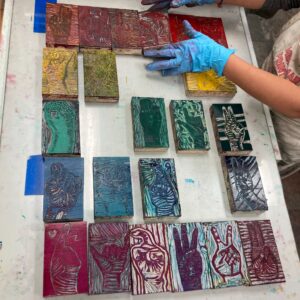 Wearing blue gloves, a creator arranges colorful hand-sign print blocks—symbols of vibrant Essex County community unity and artistry.