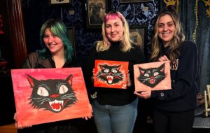 Three community members in Essex County proudly display vibrant black cat paintings, beaming amid creative energy and local art.