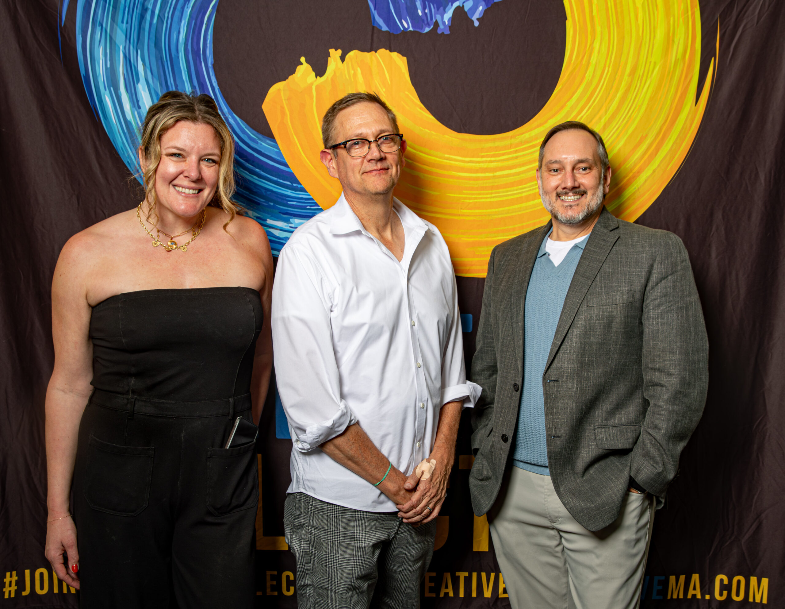 Three adults beam side by side before vibrant blue and yellow swirls, celebrating Essex County creativity and community pride.