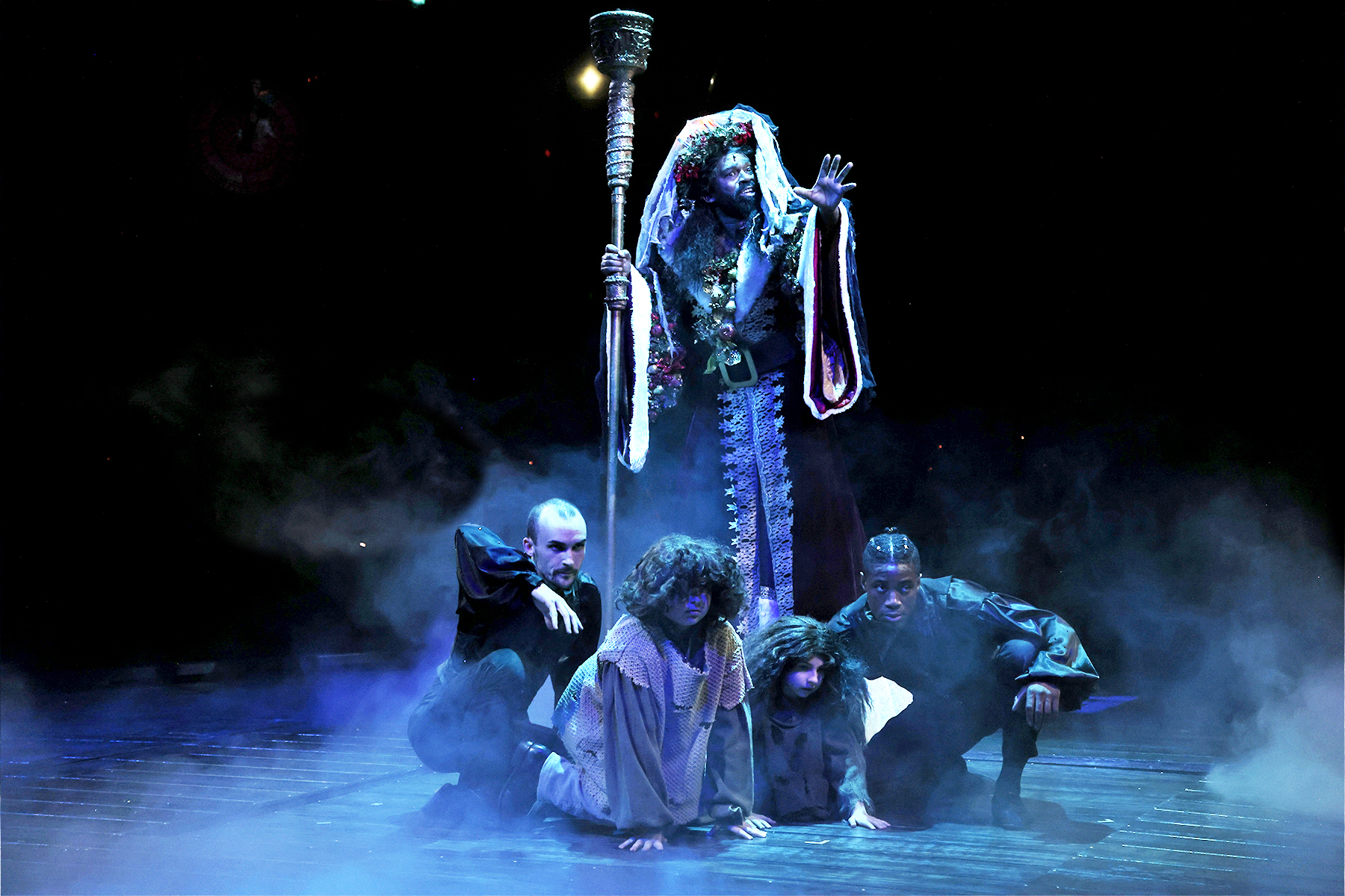 Four local performers crouch together, united in dramatic poses as a costumed figure with a staff leads; blue-lit fog fills our creative Essex County stage.