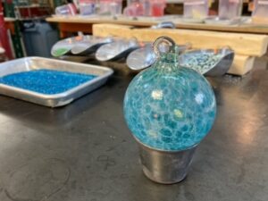 A vibrant turquoise and white glass ornament shines in a metal holder on a creative Essex County worktable, surrounded by colorful tools.