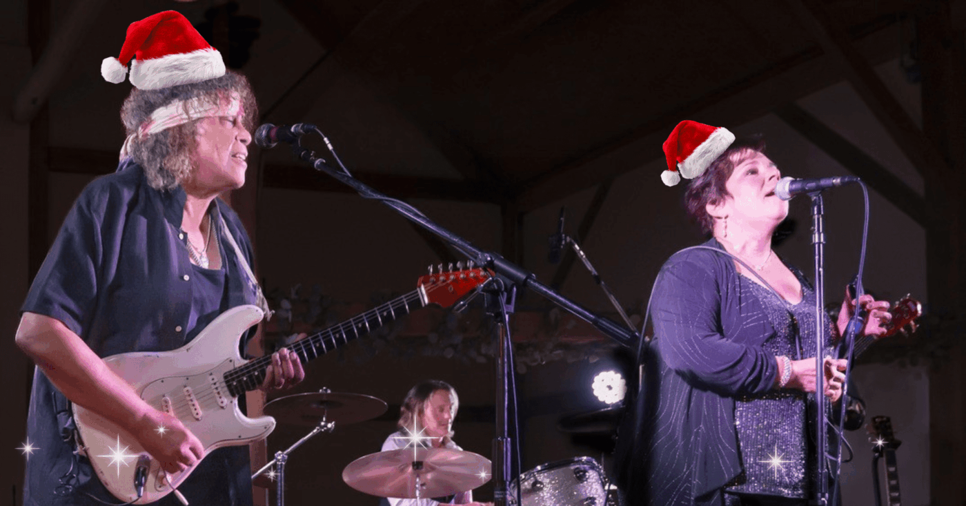 Santa-hatted women rock the stage with guitar and vocals as a drummer joins, spreading festive joy and creative spirit in Essex County.