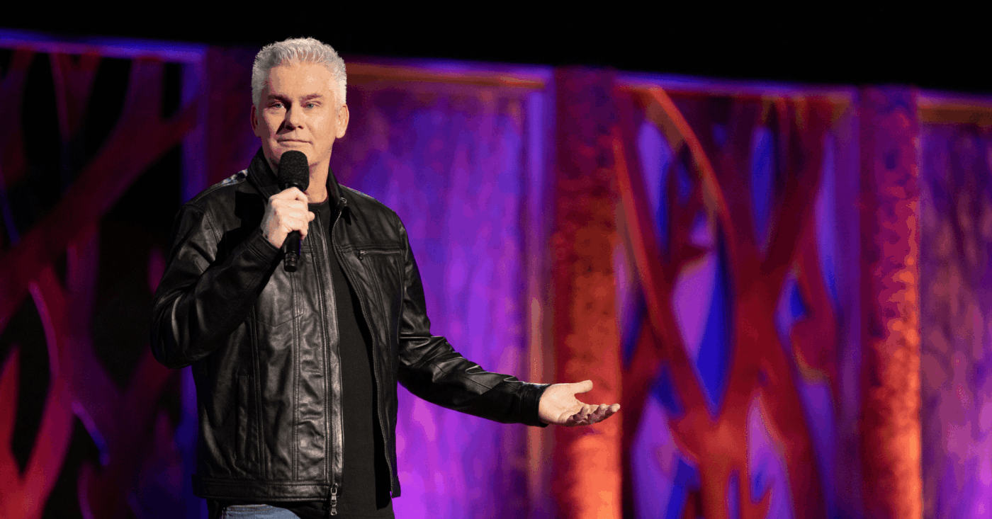 A lively man in a black leather jacket energizes the stage, mic in hand, as bold orange and purple panels spark Essex County pride.
