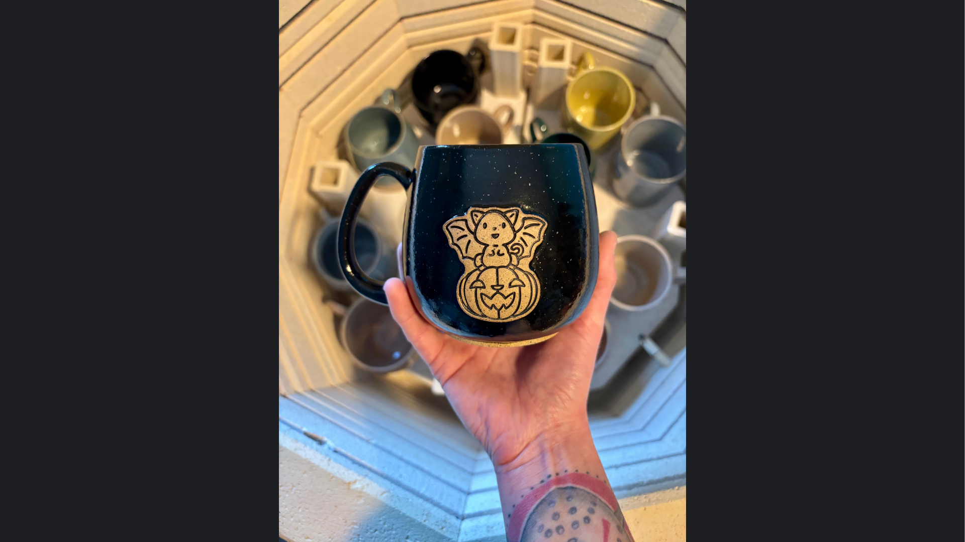 A tattooed hand proudly showcases a bold mug with a bat-winged cat; other Hippie Bean Co. mugs in the kiln celebrate Essex County artistry!.