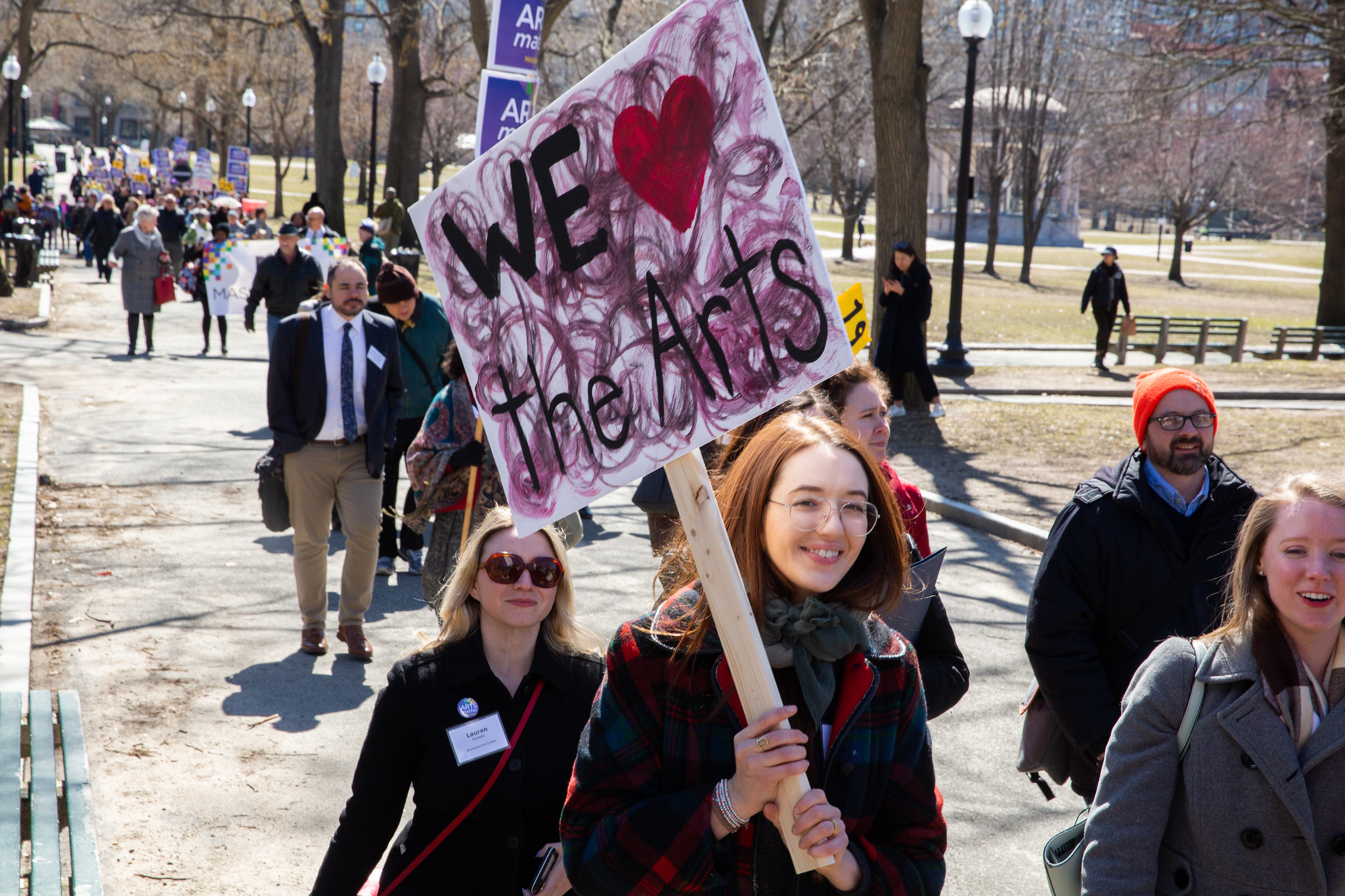 A lively crowd strolls through a sunny park, led by a woman with a "WE ♥ the Arts" sign, uniting Essex County's creative community.