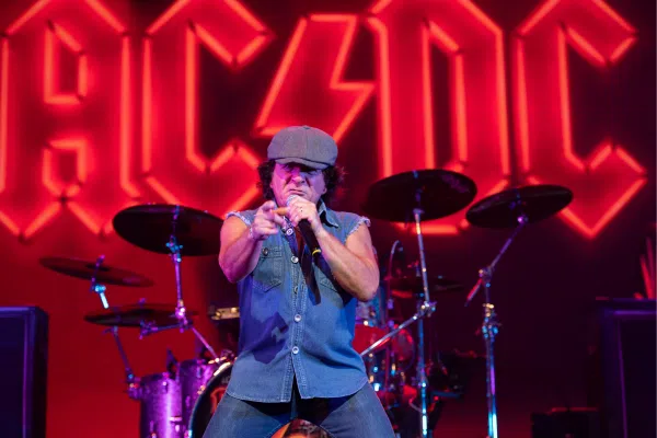 An electrifying singer in a gray cap and denim commands the stage, uniting fans as the AC/DC logo glows behind in celebration.