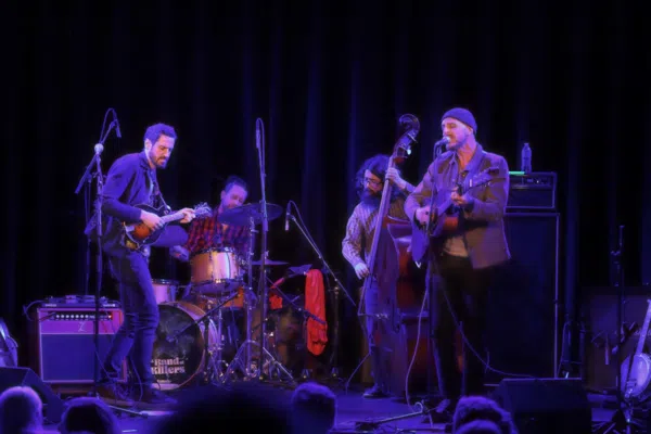 A lively four-member band electrifies the stage with guitar, mandolin, bass, and drums, uniting Essex County under vibrant blue-purple lights.
