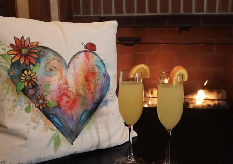 A vibrant pillow bursting with heart and flower art brightens a table with two lively mimosas, warming up community spirit by the fire.