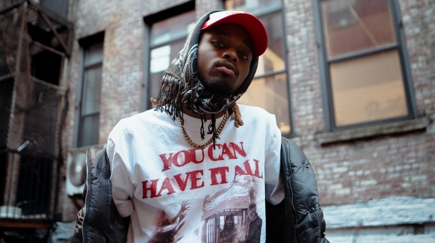 Outside a vibrant brick building, a creative soul in a red cap, bold scarf, and “YOU CAN HAVE IT ALL” tee radiates Essex County unity.