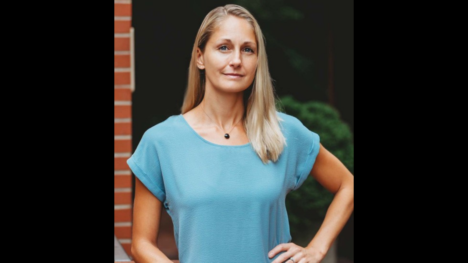 A Strangers Love’s creative spirit shines—blonde woman in blue top beams by Essex County’s brick wall and lush greenery.