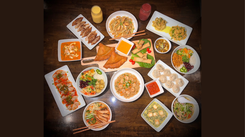 A lively spread of Asian dishes—dumplings, noodles, spring rolls, curry—fills a table, sparking community and local flavor.