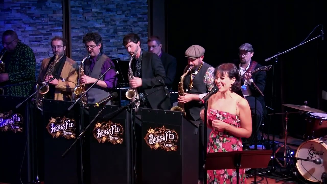 A lively jazz band with brass and woodwinds energizes the stage, as a smiling woman in a floral dress leads. “Brass Fed” stands highlight local Essex County talent connecting our community through music.