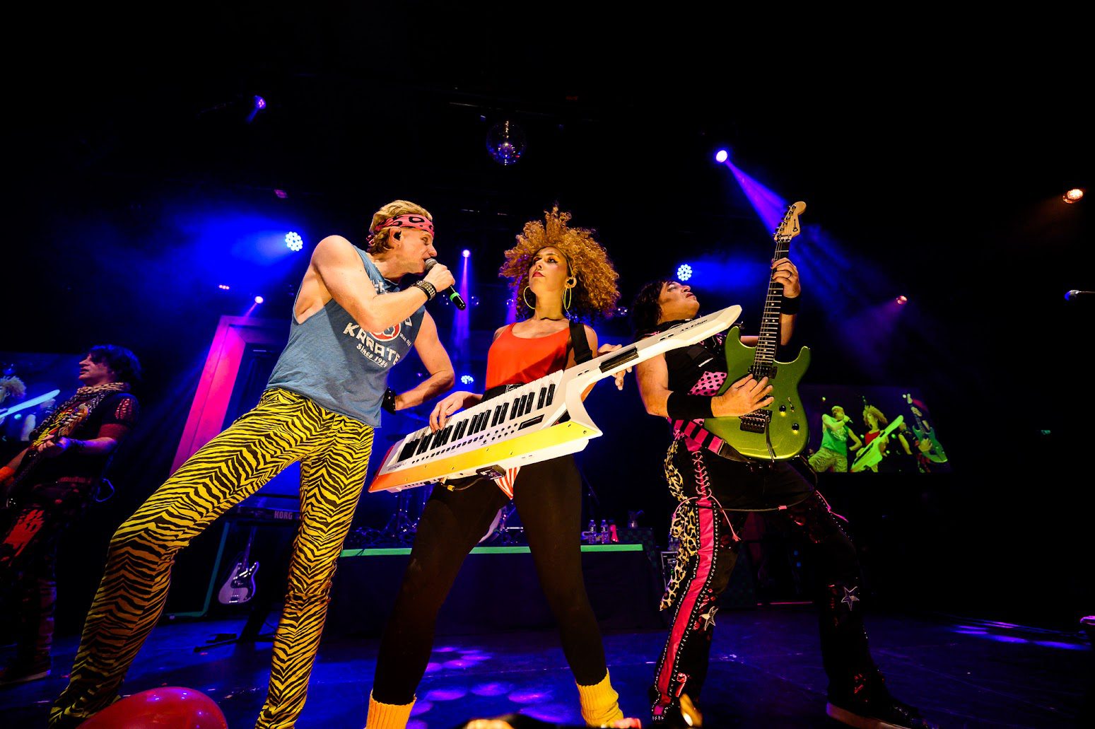 Three dynamic musicians electrify the stage in vibrant retro outfits, uniting our Essex County crowd under colorful lights.