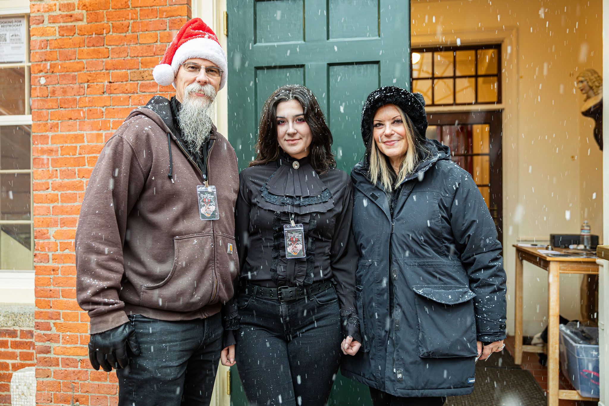 Three neighbors unite outside a brick building as snow falls, spreading holiday cheer and Essex County spirit in festive winter wear.