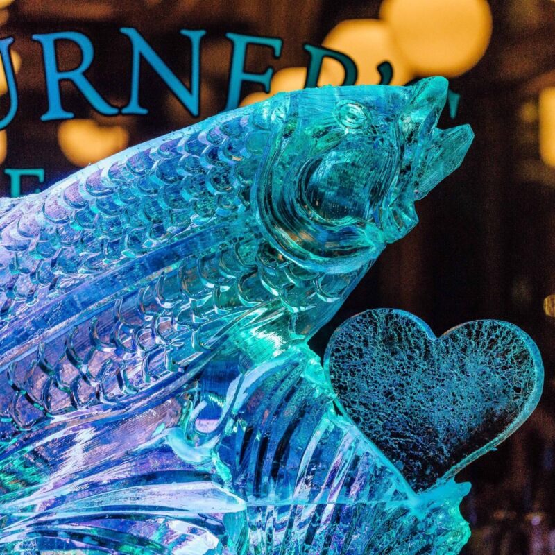 A vibrant ice fish sculpture and heart shine under blue-purple lights, celebrating Essex County’s creativity and community spirit.