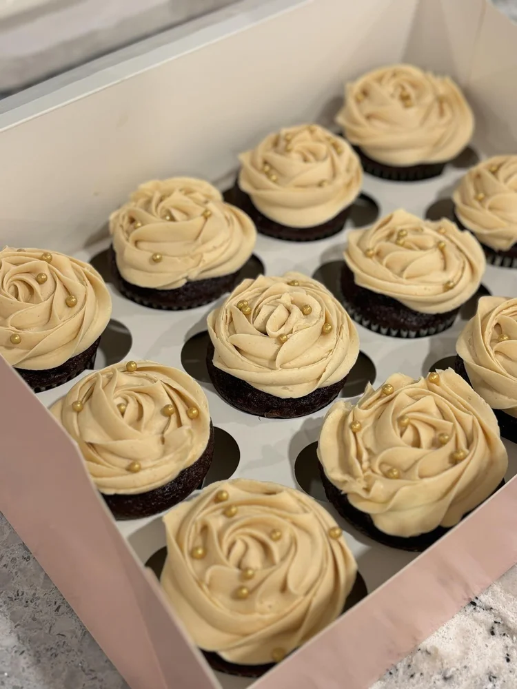 A white box of twelve chocolate cupcakes with creative rose-shaped beige frosting and gold sprinkles, baked for Essex County joy.