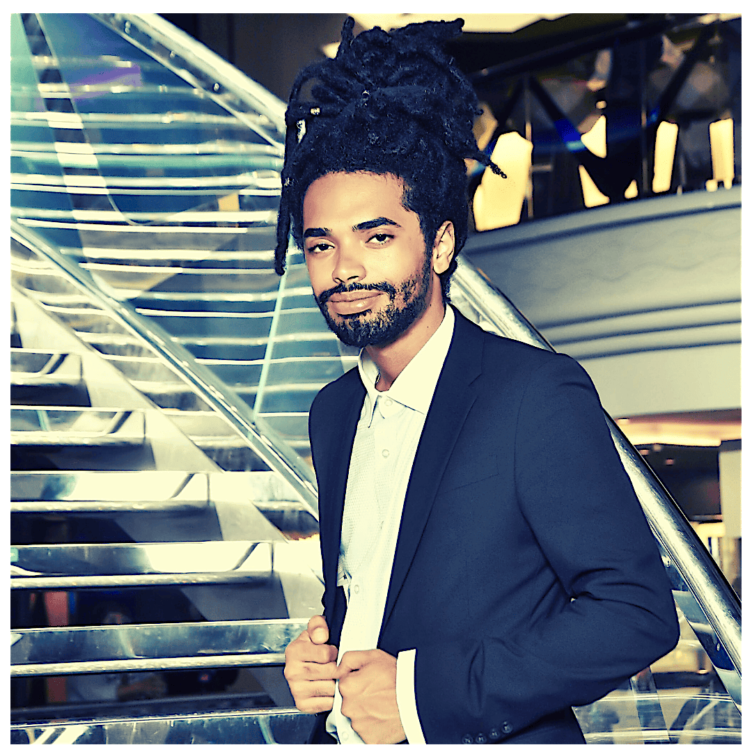 A creative leader with long dreadlocks stands confidently by a glass staircase, embodying the vibrant spirit of Essex County.