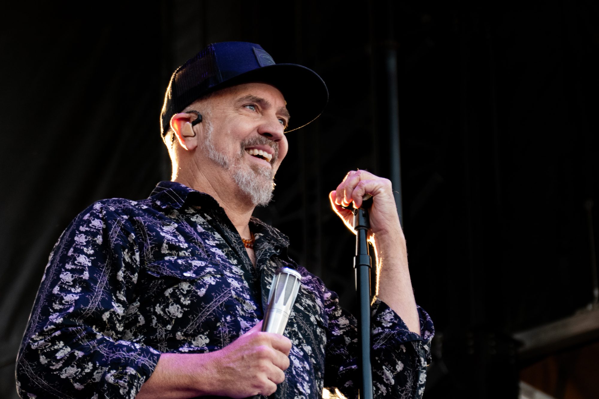Beaming on stage at an Essex County festival, a gray-bearded man in a patterned shirt energizes the crowd with lively music and local spirit.