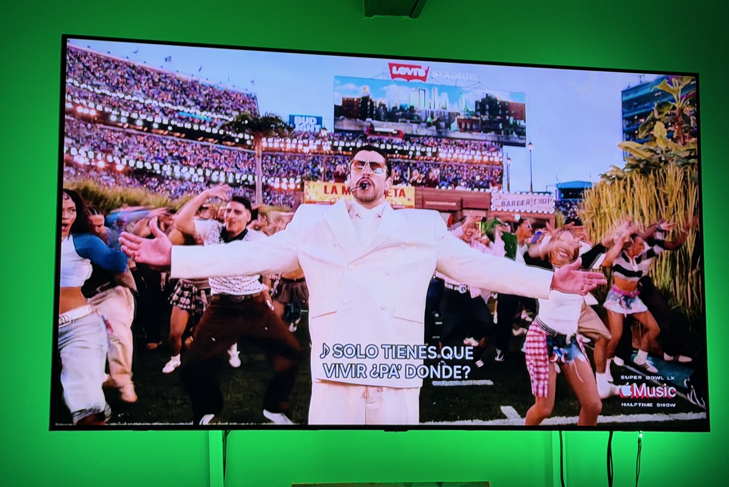 A lively TV broadcast captures a dazzling performer in white leading dancers on a stadium stage, vibrant Spanish subtitles, and green glow uniting the community. Bad Bunny Super Bowl