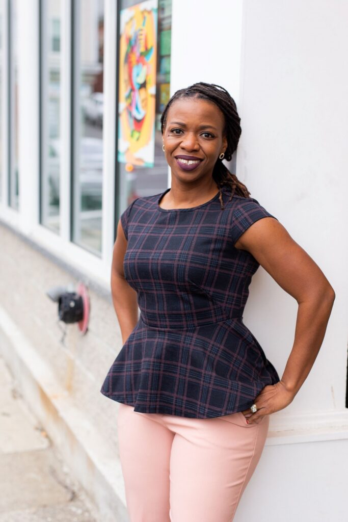 A vibrant woman beams with Black Excellence outdoors in Essex County, leaning on a white wall near lively art and bustling community life.