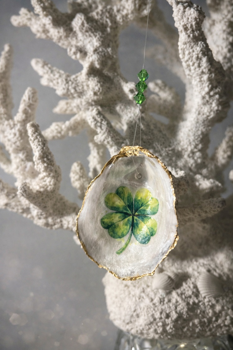 A vibrant oyster shell ornament with a green four-leaf clover, handcrafted by Essex County makers, hangs with beads before white coral.