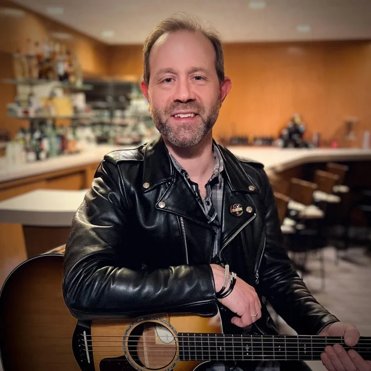 A musician in a black leather jacket livens up a cozy Salem bar, acoustic guitar in hand—celebrating Essex County’s creative spirit.