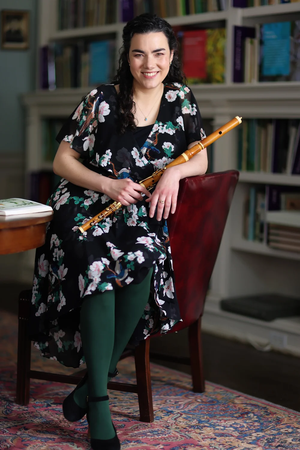 A woman with dark curly hair, wearing a black floral dress and green tights, sits on a red chair holding a wooden flute. She is smiling in a room lined with bookshelves, representing Essex County Artists supported by Massachusetts Cultural Council Grants.