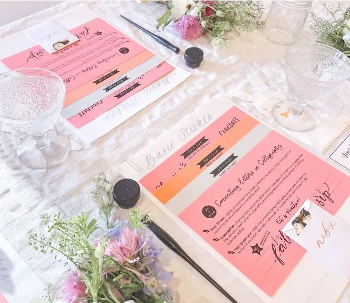 A lively beginner calligraphy table in Essex County, buzzing with pink guides, shared tools, and flowers sparking creativity together.