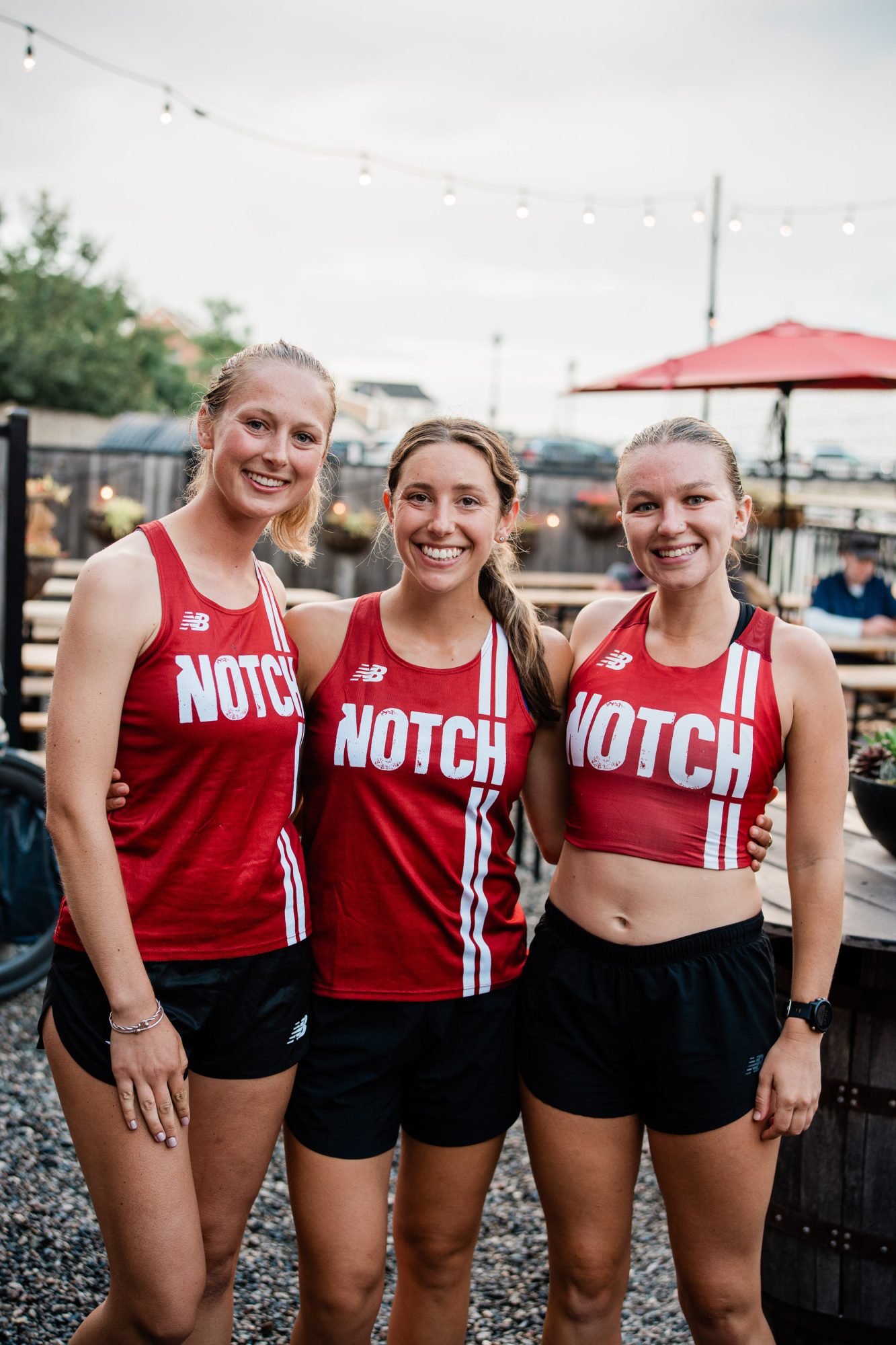 Three women wearing matching red "NOTCH" athletic tank tops and black shorts stand side by side outdoors, smiling at the camera. There are string lights and patio seating in the background.