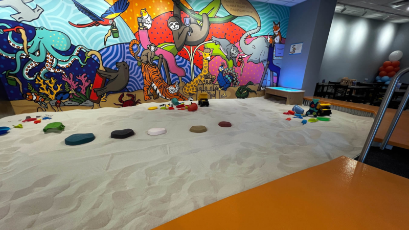 Vibrant indoor sandbox filled with colorful toys and plastic rocks, lively mural, and festive balloons—where Essex County families connect and create.