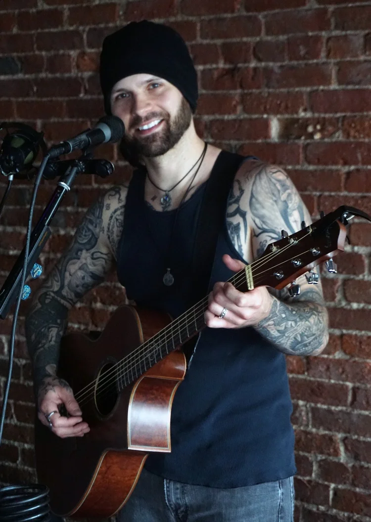 A tattooed musician in a black beanie and tank top beams while strumming guitar, energizing our Essex County community with live John & Jen music.