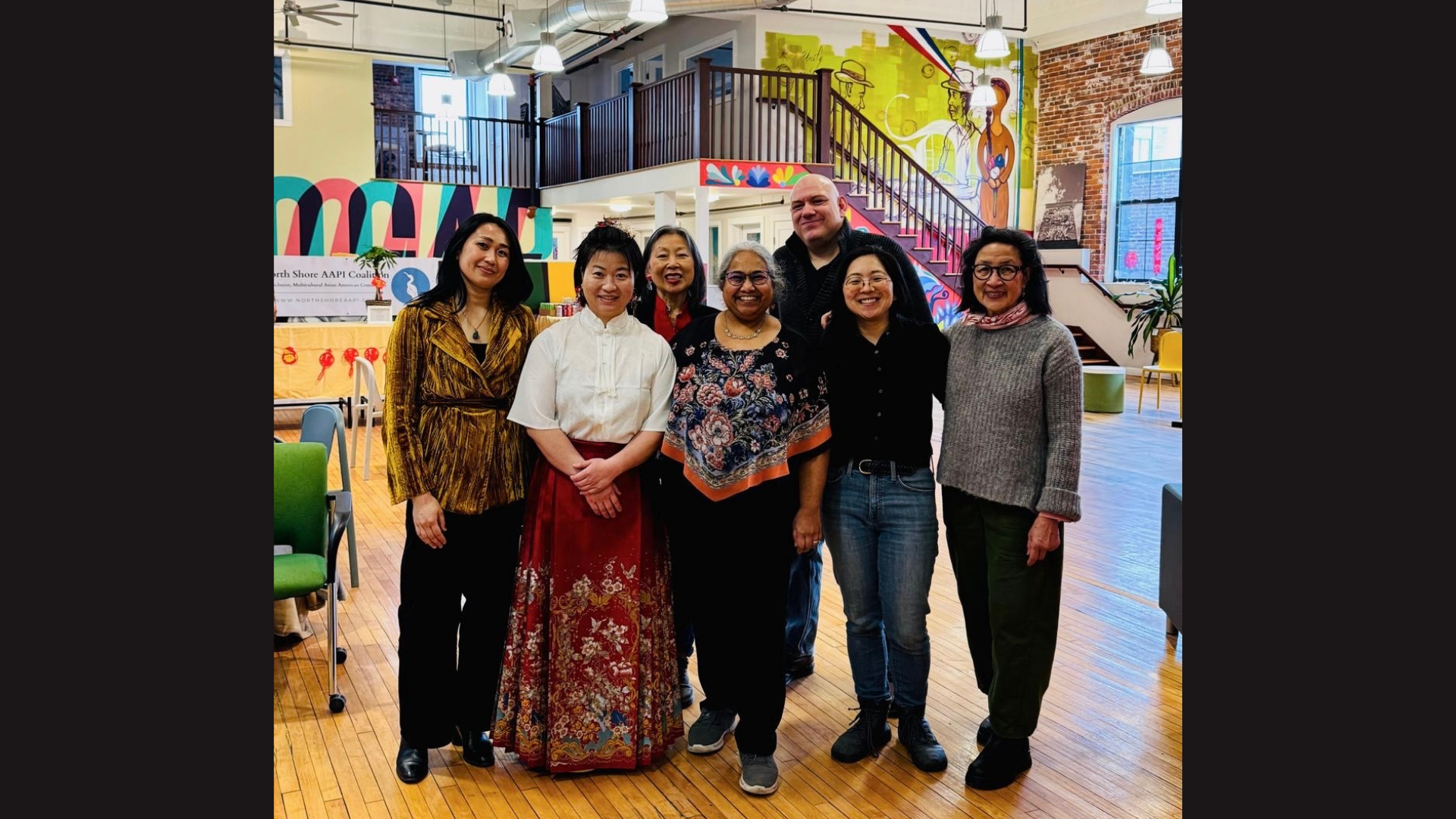 Seven people stand together and smile for a group photo in a brightly lit, colorful indoor space at Meet the Member: ECS Wellness, with art on the walls, exposed brick, wooden floors, and an open staircase in the background.