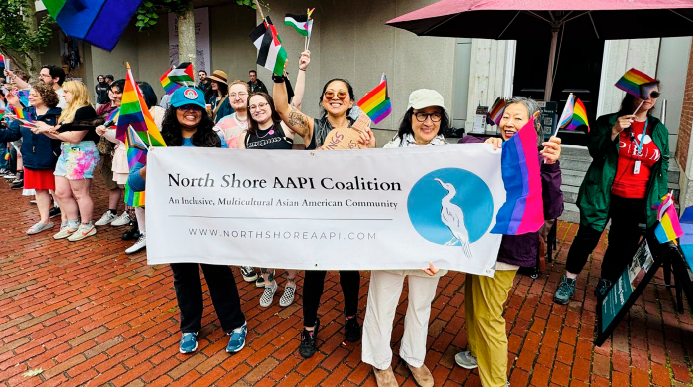 A group stands on a brick sidewalk, proudly holding a North Shore AAPI Coalition banner and waving rainbow flags at an outdoor event. Others in the background join in, smiling and showing their support with more flags.