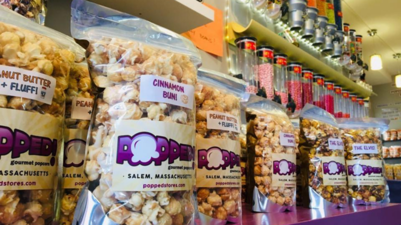 Bags of flavored popcorn, including Cinnamon Bun and Peanut Butter + Fluff, are lined up on a purple counter. Candy dispensers filled with colorful sweets are mounted on the wall in the background.