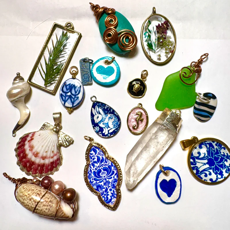 A collection of handmade jewelry pendants arranged on a white surface, featuring custom charms crafted from crystals, seashells, painted ceramics, wire wrapping, pressed flowers, and different colored stones.