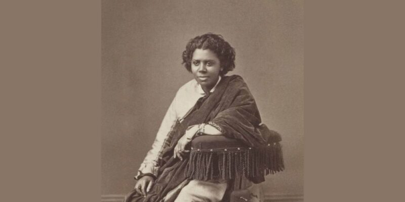 A creative woman with lively short curls, in a light dress and bold fringed shawl, sits confidently—her spirit brightening Essex County.