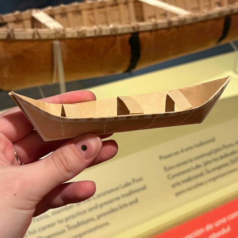 A hand displays a crafted cardboard canoe model, with larger canoe and exhibit behind, celebrating Essex County’s creative community spirit.