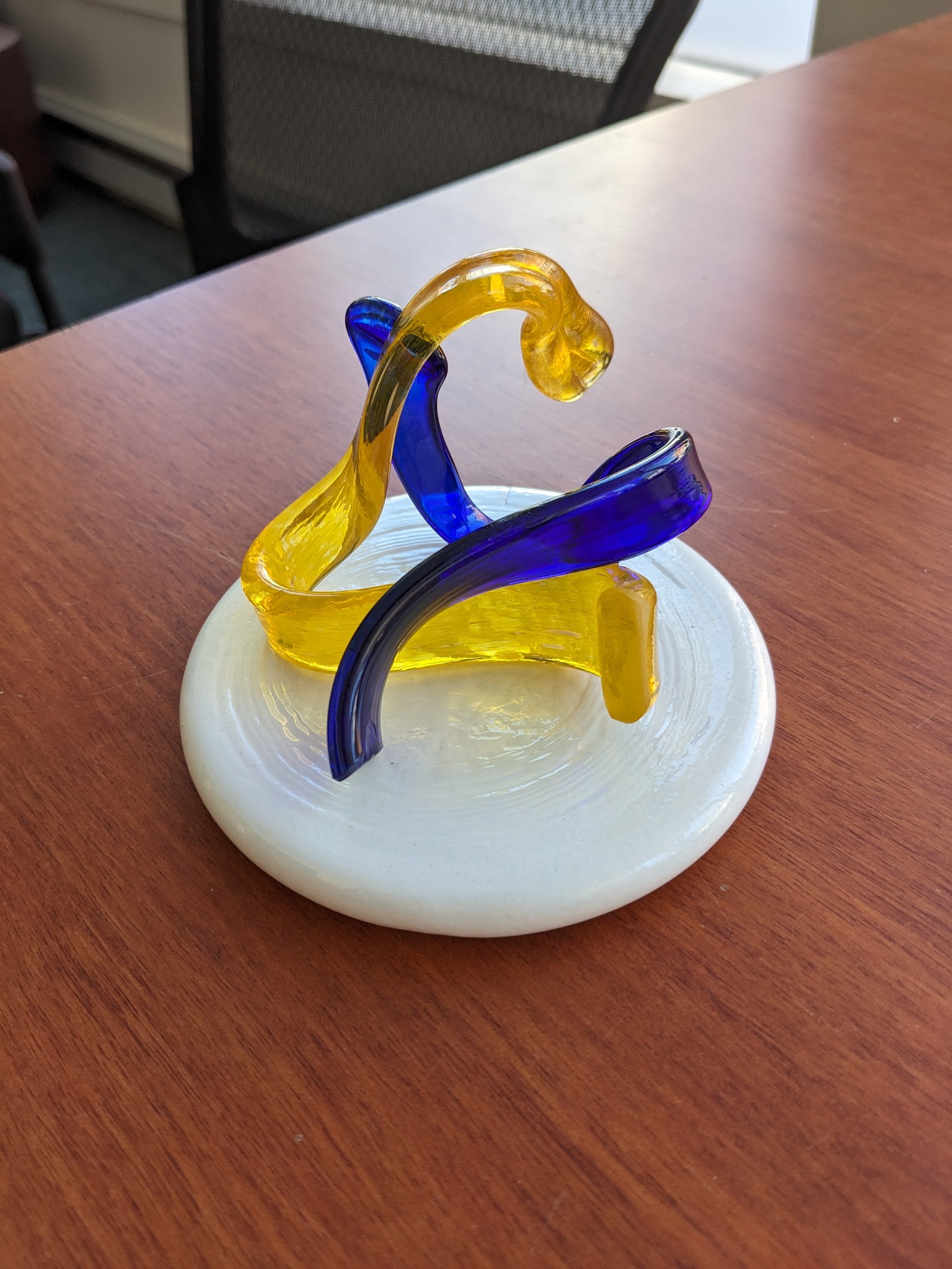 A lively abstract sculpture with swirling yellow and blue glass beams creativity, uniting Essex County on a bright white base.