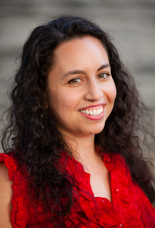 A woman with long, curly dark hair smiles at the camera. She is wearing a sleeveless red top with ruffle details. The blurred gray background highlights her joy—reflecting the spirit of Essex County artists and Massachusetts Cultural Council grants 2026.