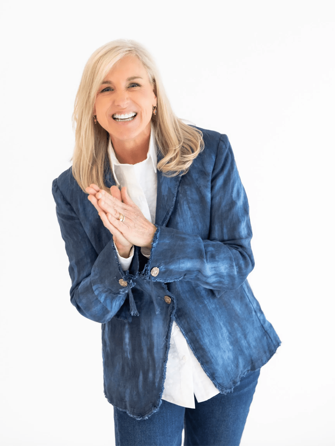 A woman with long blonde hair wearing a blue blazer, white shirt, and blue jeans stands against a white background, smiling and clasping her hands together.