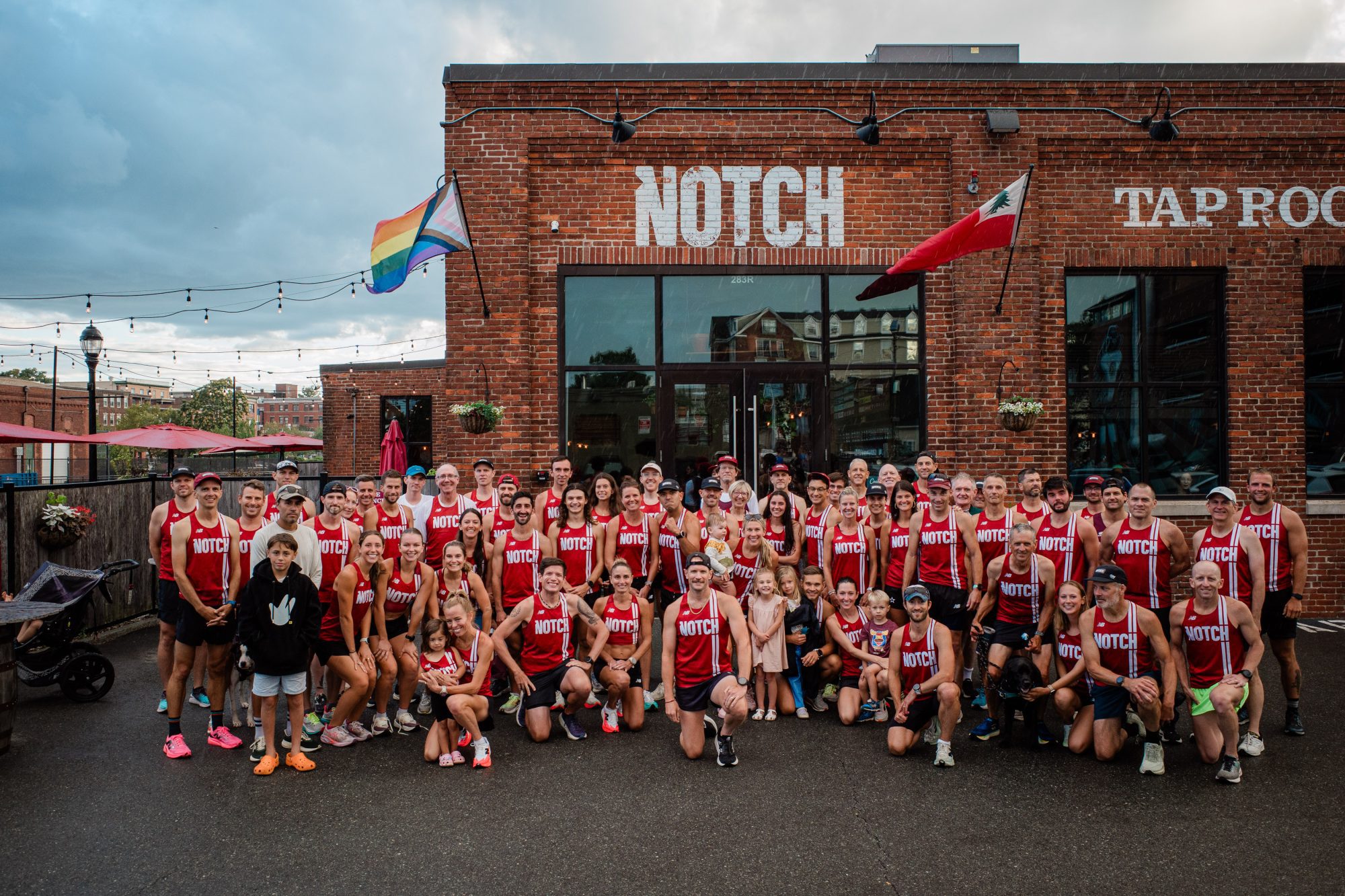A large group of people wearing matching red tank tops with “NOTCH” printed on them pose for a group photo outside a brick building with “NOTCH TAP ROOM” on the facade. Two flags hang near the entrance.
