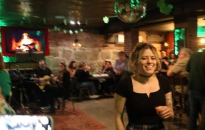 A smiling woman in a black top stands amid lively conversation at an Essex County Irish pub, glowing with community and local spirit.