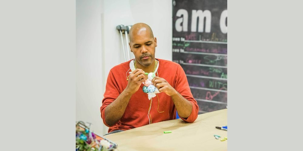A man wearing a red sweater sits at a table and works with yarn in his hands. A container of craft supplies is on the table, and a chalkboard wall is visible in the background.