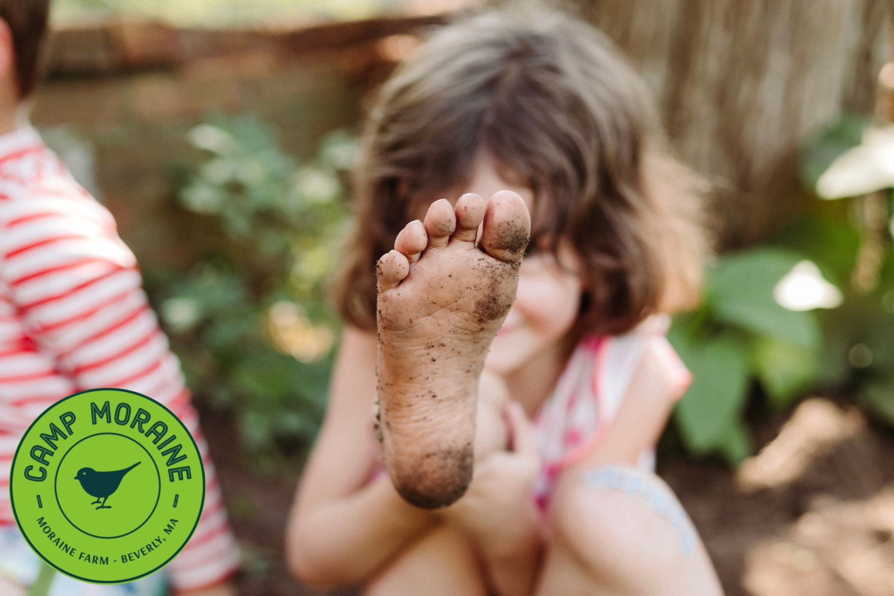 A joyful child with light brown hair shows a muddy foot to the camera, surrounded by greenery—Camp Moraine invites Essex County kids to connect and create!.