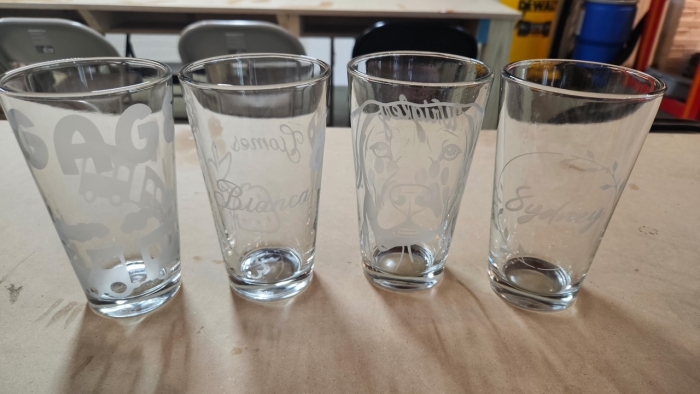 Four etched pint glasses showcase local artistry on a table in Essex County, exuding creative community spirit; chairs behind hint at gatherings.