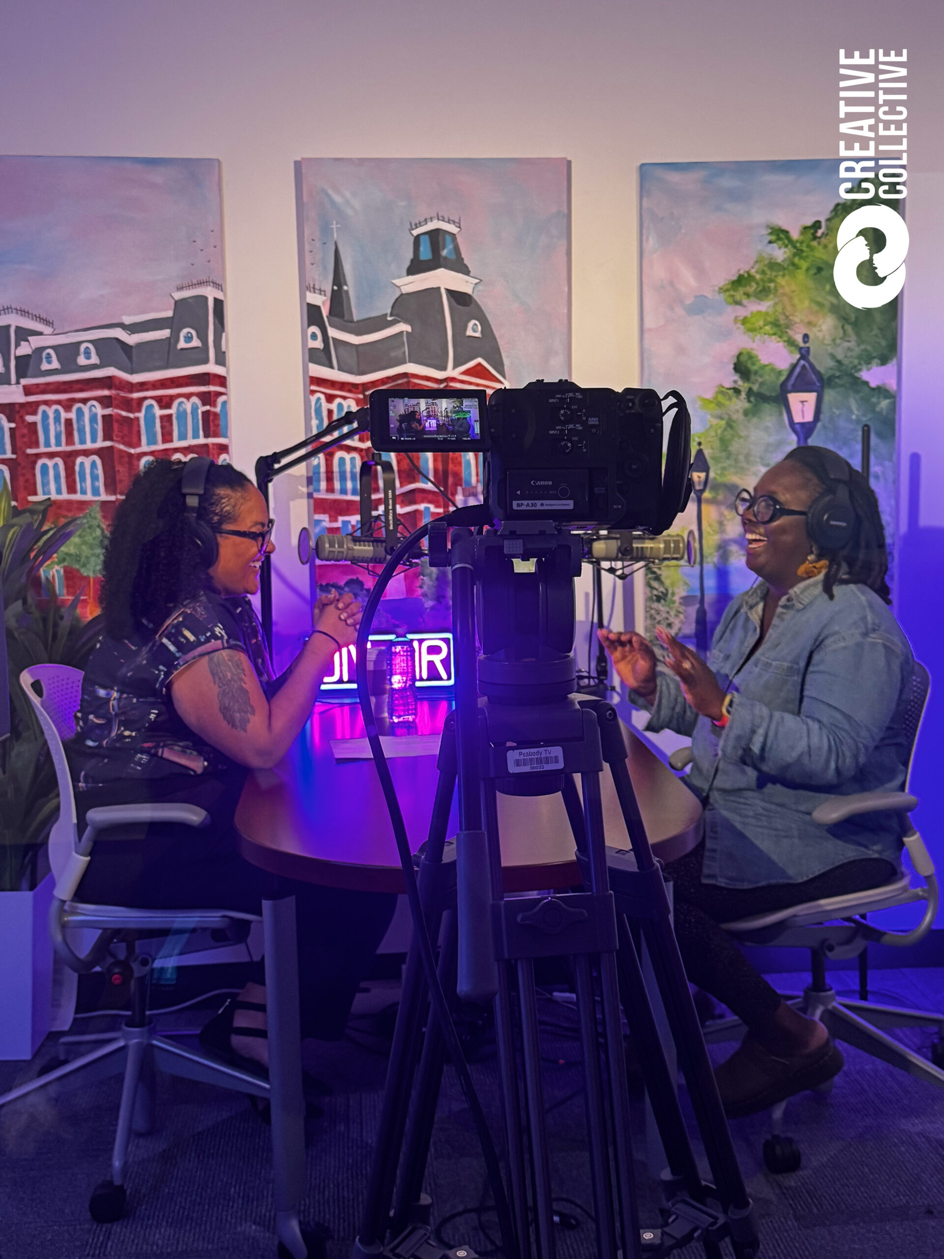 Two women podcast at a lively table, surrounded by bright art and a "Creative Collective" logo, energizing our Essex County scene.