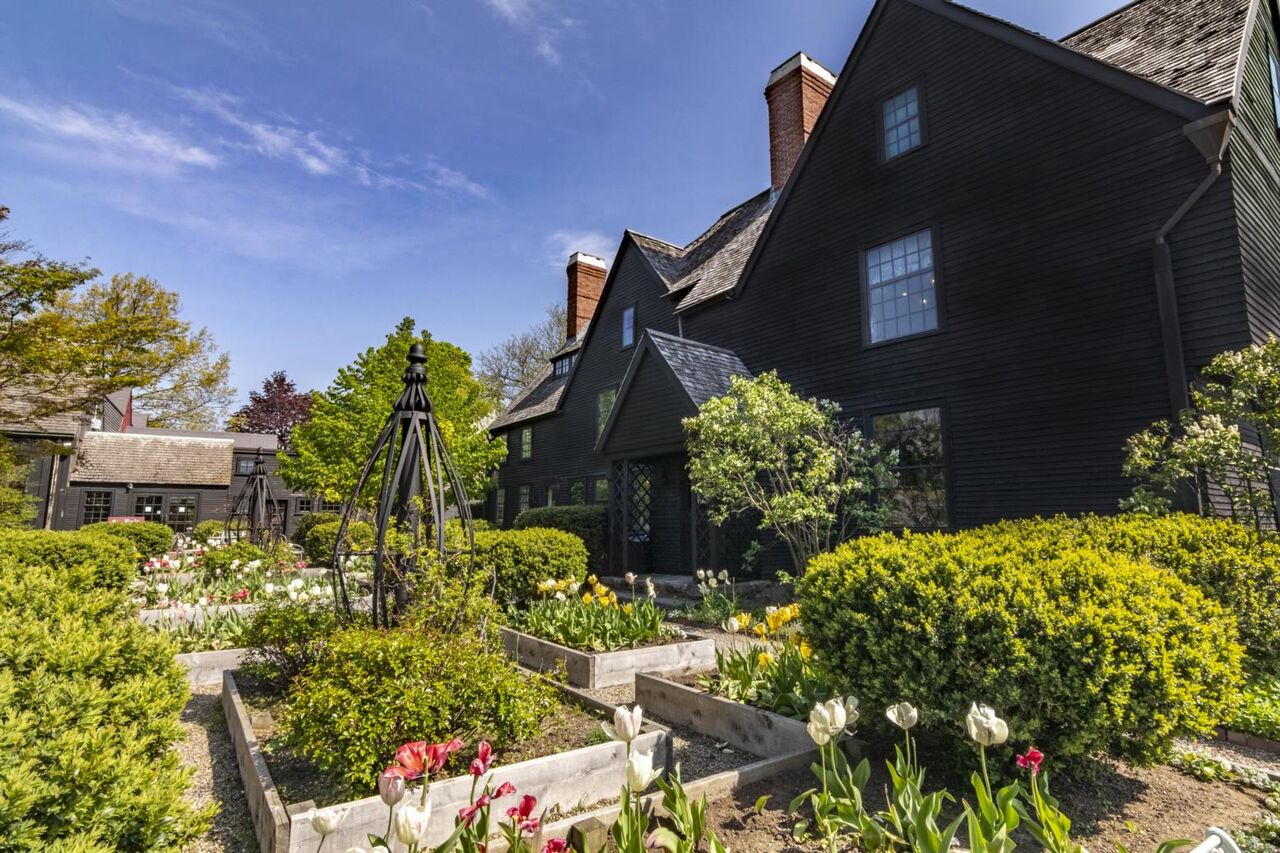 A striking black house with steep roofs rises behind a lively Essex County garden, where local blooms and creative touches unite the community.